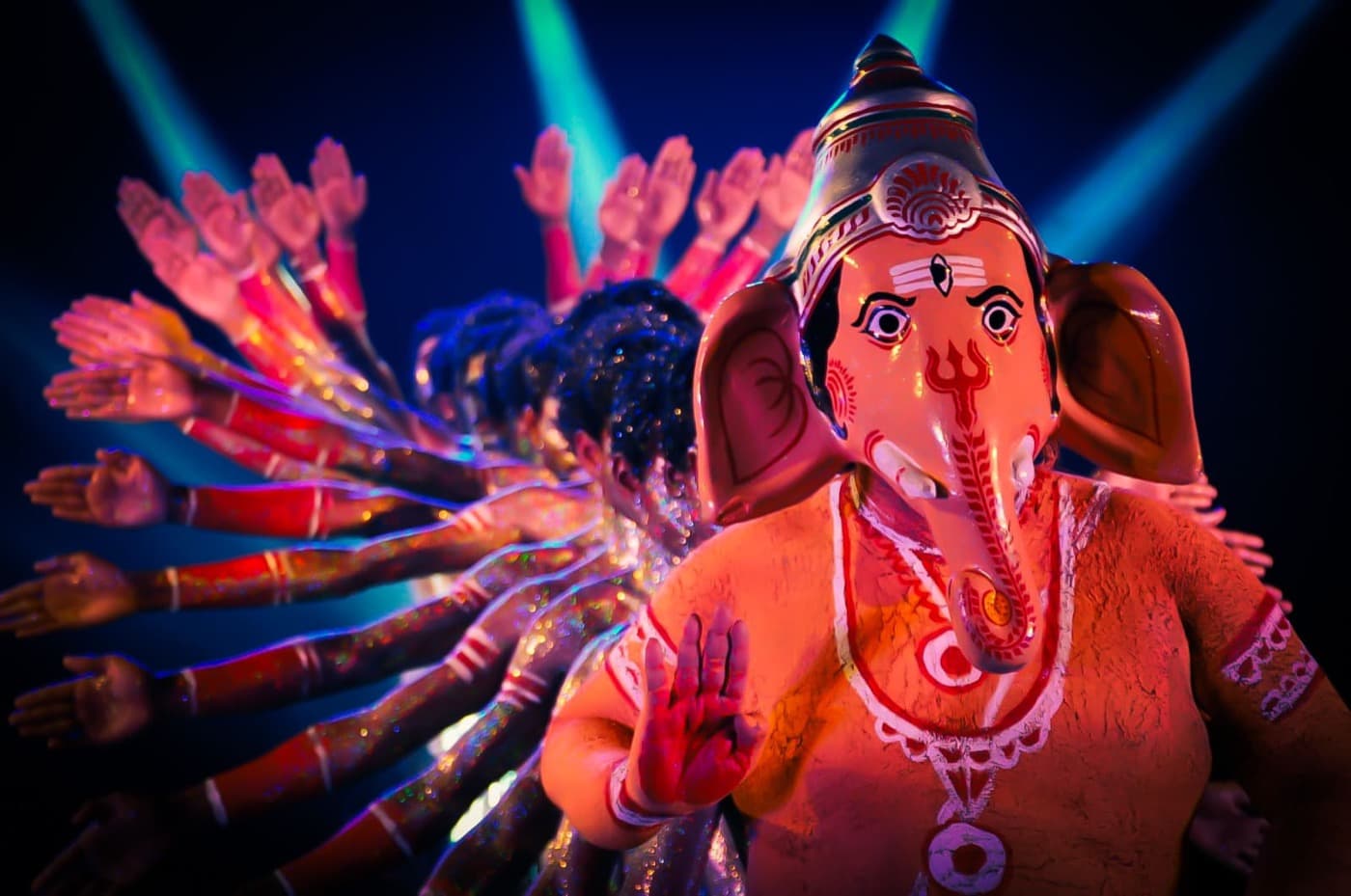 Prince Dance Group performer in an ornate elephant-masked costume backed by an ensemble in metallic body paint with arms raised, under blue and pink stage lighting