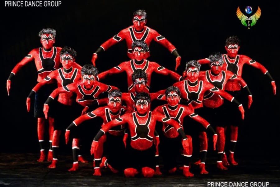 Prince Dance Group ensemble in red and black body paint forming a human pyramid on a dark stage